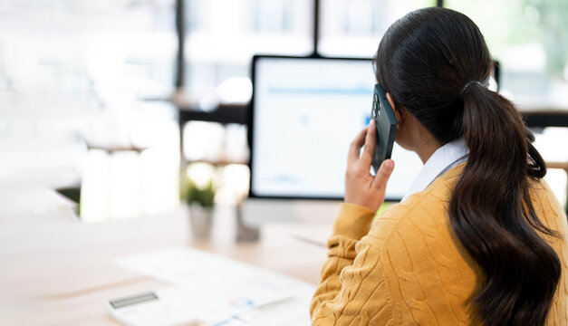 Back view of Asian woman with ponytail in yellow sweater talking on smartphone in bright office.