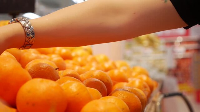 A woman chooses mandarins among a large pile of citrus fruits displayed across a supermarket fruit counter. Fresh citrus.