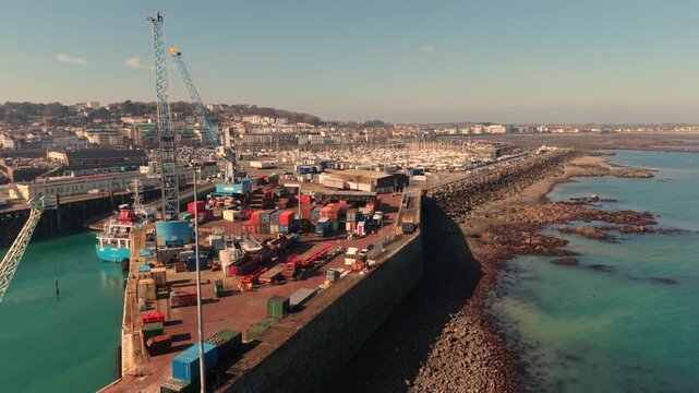 St Peter Port Harbour Guernsey.Low drone footage over working docks with QEII Marina,town and Belle Greve Bay behind and seabirds flying by in late afternoon sun with calm clear blue water