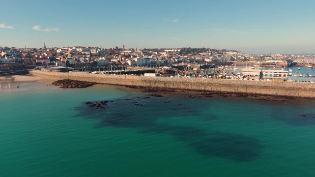 Havelet Bay St Peter Port Guernsey.Reveal of the bay,seawall and beach with clear blue water and the harbour and town seafront as its backdrop in late afternoon sun with cloudless sky