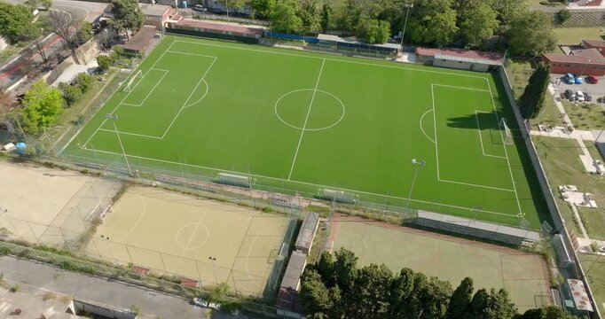 Aerial perspective of a lush green football pitch with white markings, situated within a sports facility. The empty field is bordered by trees and adjacent courts, perfect for team sports and training