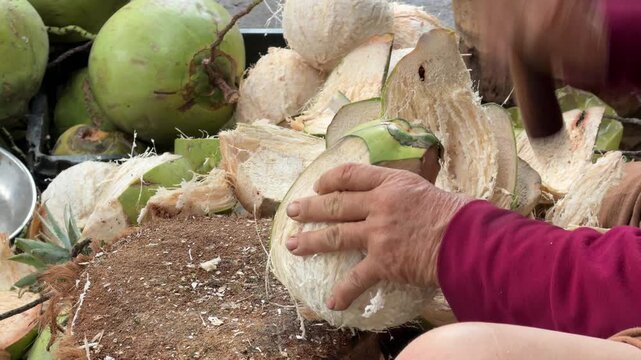 Market vendor chopping and peeling a coconut with a machete on a wooden block surrounded by coconuts at a street market stall. Coconut preparation, street market.