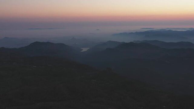 Yercaud's layered mountain ridges emerge through morning mist under pastel dawn sky in Tamil Nadu, India. Slow drone shot captures the hill station's elevated terrain with fog settling in valleys.