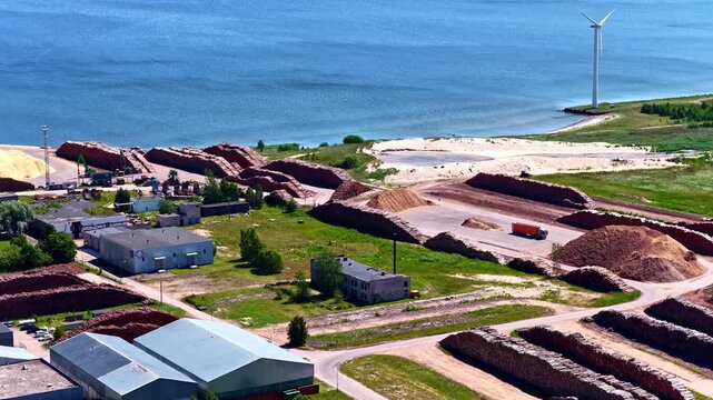 Aerial flyover of a coastal timber processing factory in Latvia with large raw material mounds, warehouses, a red truck, and wind turbines standing on a sandy Baltic Sea shoreline