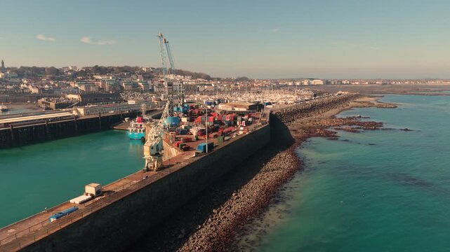 St Peter Port Harbour Guernsey.Low drone footage along the docks and seawall at the entrance to the harbour showing working docks and ferry berth in late afternoon sun with calm clear blue water