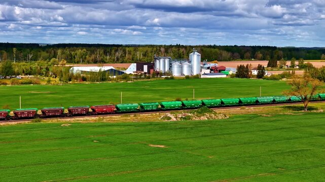Aerial view of long stationary freight train with green and red hopper wagons alongside grain silos and agricultural facility amid lush green Latvian spring countryside