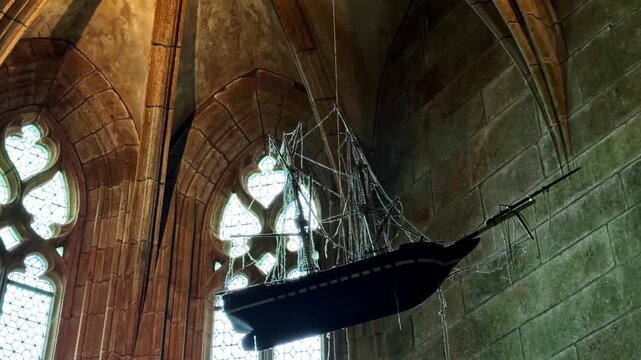A detailed ship model hangs inside the historic Mont-Saint-Michel Abbey, framed by Gothic arches and stained glass. Ideal for use in historical documentaries or architectural studies.
