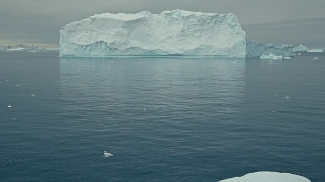 Massive flat-topped tabular iceberg with sheer cliff walls floats in calm grey-blue Antarctic ocean waters surrounded by smaller ice fragments