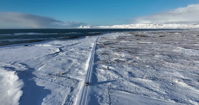 Aerial view of a lone 4x4 driving toward Strandarkirkja across Iceland&rsquo;s snowy coastal plain, with the Atlantic shoreline and vast winter landscape stretching ahead.