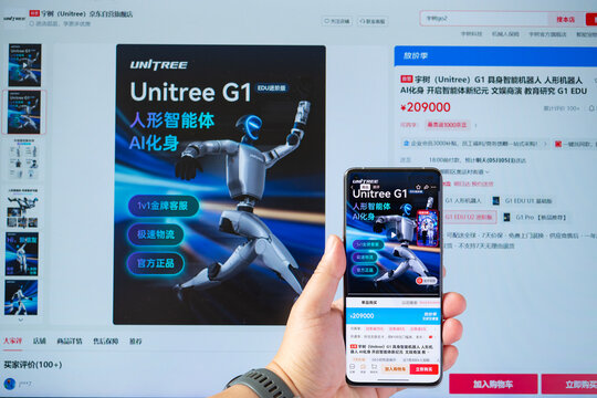 ZHONGSHAN China-May 4 2026: hand holding a smartphone displaying the Unitree G1 humanoid robot product page while a monitor shows the same product listed on JD.com.