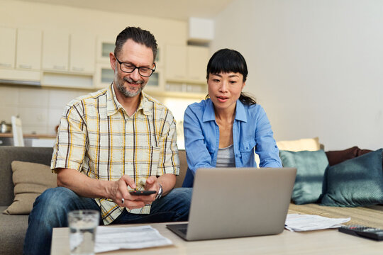Young couple checking bills and calculating expenses using a laptop at home, debt taxes concept, worried married elderly couple