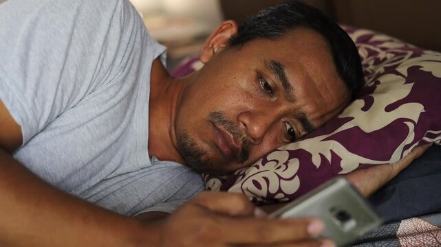 Depressed Asian man lies on the bed and scrolling mobile phone feed. Concept of depression or fatigue. Disappointed sleepy male starring at mobile phone screen and thinking.