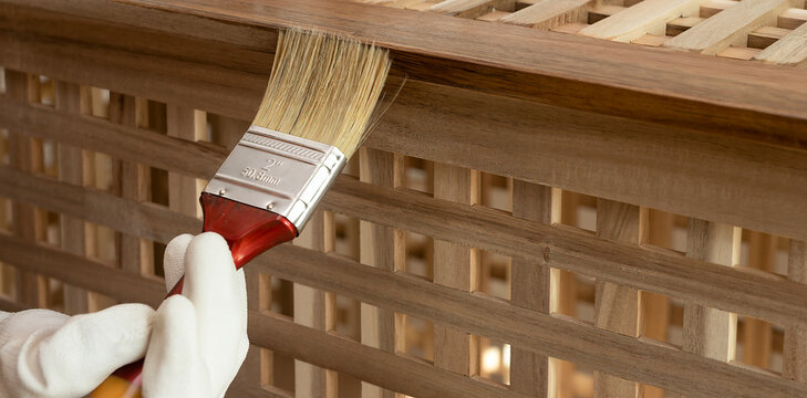 Paintbrush applies finish to an acacia wood lattice table. Gloved hand coats the slatted surface during furniture maintenance. DIY woodwork and home improvement concept..