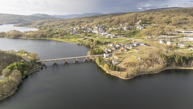 Aerial view of the stone arched bridge crossing the Encoro de Prada reservoir with the village of A Veiga nestled on the green hillside under a soft sky in A Veiga, Ourense, Spain.