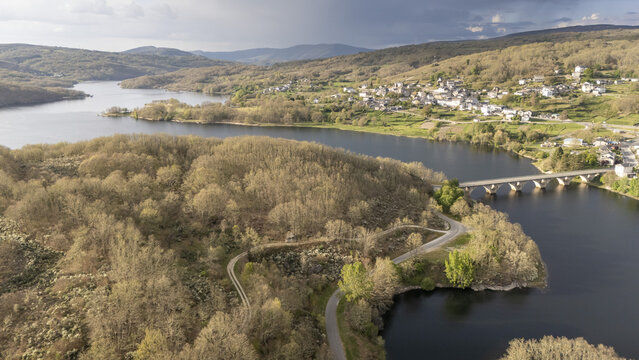 Aerial view of Encoro de Prada reservoir with a stone bridge and a hillside village surrounded by dense forests in A Veiga, Ourense, Spain.