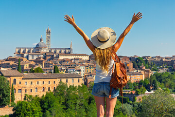 Naklejka premium Happy young woman traveler with leather backpack and straw hat standing with arms raised and enjoying the scenic skyline view of siena, italy