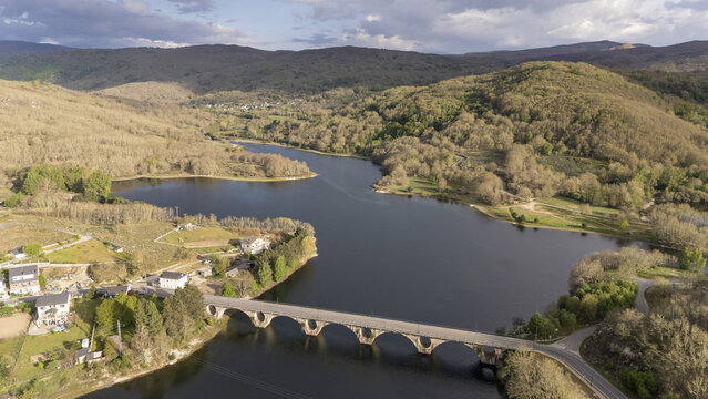 Aerial view of Encoro de Prada reservoir with a stone arch bridge crossing the water, surrounded by lush green hills and rural houses in A Veiga, Ourense, Spain.