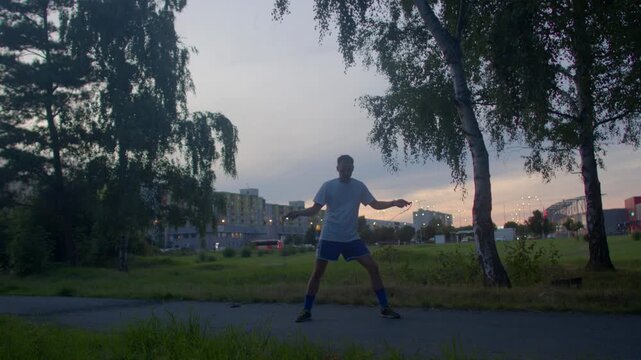 man exercising city park dusk, rapid jump rope session under birch trees with urban skyline backdrop, evening glow, focused posture, rhythmic footwork and high energy cardio training
