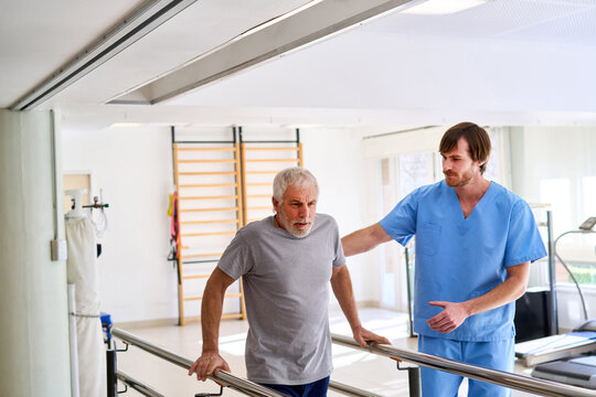 Senior Man Walking With Physiotherapist Assistance On Parallel Bars In Rehab Center For Injury Recovery And Strength Training Indoors