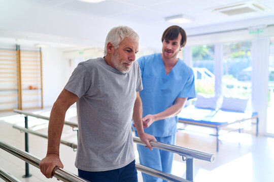 Senior Man Walking With Parallel Bars Assisted By Physiotherapist In Rehab Center For Injury Recovery And Rehabilitation Indoors