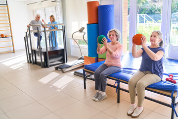 Naklejka premium Senior Women Doing Ball Exercises On Bench In Rehab Center With Physiotherapist Assisting Man On Walking Aid For Rehabilitation Indoors