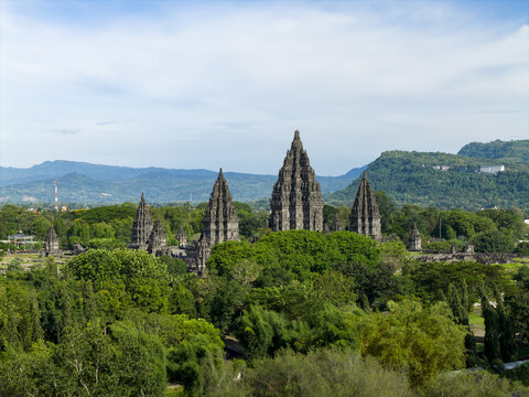 Aerial view of Prambanan Temple complex with its iconic stone spires rising above lush tropical greenery and distant mountains under a bright sky Prambanan, Daerah Istimewa Yogya, Indonesia.