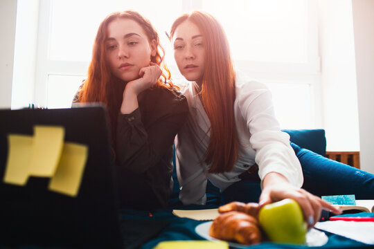 Two red-haired students study at home or prepare for exams. Young women doing homework in a dormitory bed near the window. There are notebooks, food books, a tablet and laptop and documents