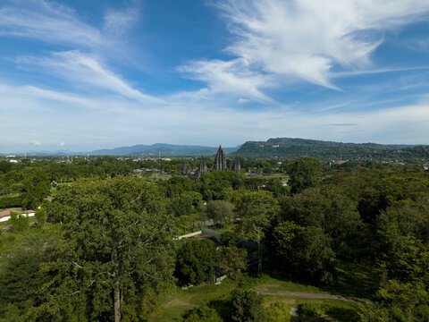 Aerial view of the ancient Prambanan Temple complex nestled among lush tropical forests and rolling hills under a bright blue sky with wispy clouds Prambanan, Daerah Istimewa Yogya, Indonesia.
