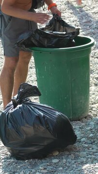 Volunteer beach clean-up, person placing a full black garbage bag into a green trash can on a sunny pebble shore. Vertical video