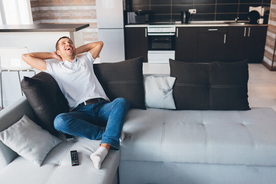 Young man watch tv in his own apartment. Sit on couch and laughing out loud. Loook up and hold hands under head. Relax, rest and enjoy. Alone in room. Daylight.