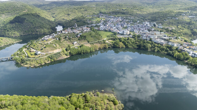 Aerial view of the town of Viana do Bolo surrounded by lush green hills and the calm waters of the Bibei river with cloud reflections Viana do Bolo, Province of Ourense, Spain.