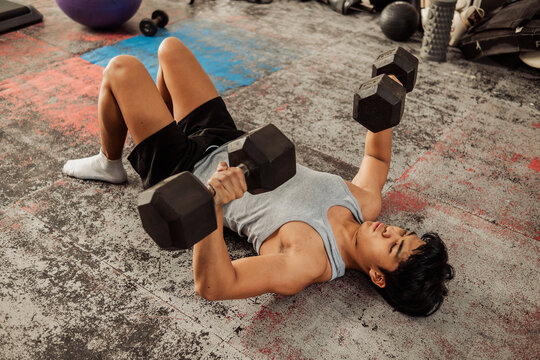 Young Asian man does dumbbell floor press at the gym focusing on chest, triceps, and shoulder strength using controlled resistance training on the floor