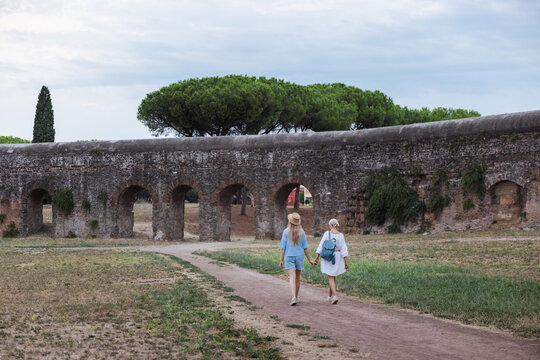 Mother and teenage daughter holding hands walking toward ancient Roman aqueduct arches in Parco degli Acquedotti Rome enjoying travel in Italy