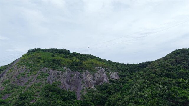 Sugarloaf Mountain cable car, gliding over dense tropical forest under a moody, grey sky in Rio de Janeiro, Brazil. Forward upward tilted aerial approach.