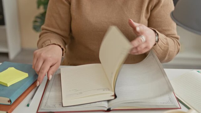 Woman pinches nose and closes eyes over open notebook at desk with books, sticky notes, cup and lamp in building; fatigue.