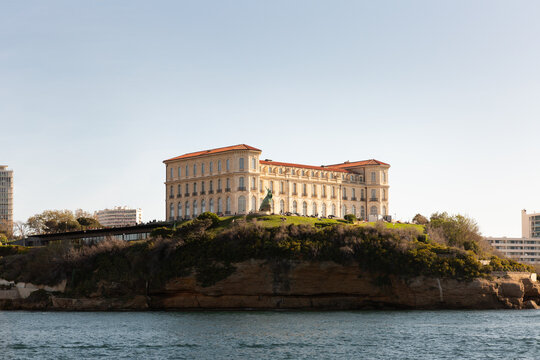 Palais du Pharo &agrave; Marseille vue depuis la mer, rocher avec de la verdure en contre-bas, mer au premier plan, lumi&egrave;re douce et ensoleill&eacute;e, ciel bleu