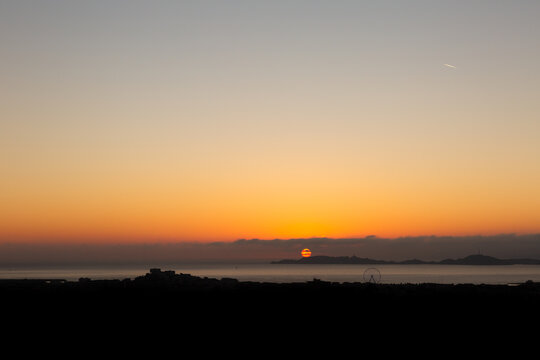 Coucher de soleil sur la mer &agrave; Marseille, soleil orange flamboyant se couchant derri&egrave;re les &icirc;les du Frioul, ville dans l'obscurit&eacute; au premier plan, grand ciel avec un beau d&eacute;grad&eacute; bleu orang&eacute;