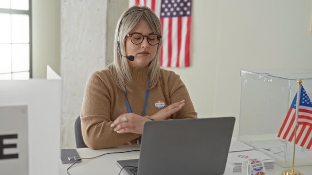 Woman wearing headset and glasses types on laptop at polling station table with clear ballot box and i voted sticker in building; civic duty friendly.