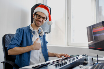 Thumb up. Merry Christmas and a happy new year! Composer hands on piano keys in recording studio. music production technology, man in christmas hat is working on pianino and computer keyboard on desk. © Vlad