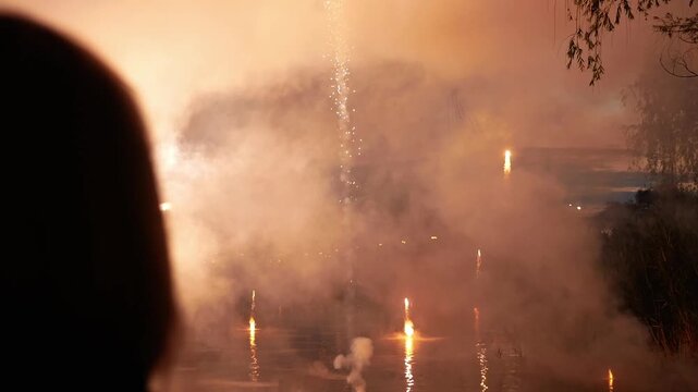 Silhouette of a woman photographing fireworks in the night sky. Beautiful fireworks in honor of the holiday. A woman takes pictures on her phone as golden sparks fly into the sky.