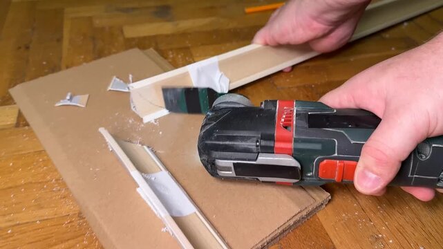 Hands use a cordless power drill to fasten a small wooden frame on a cardboard work surface, indoor diy repair scene with focused practical mood