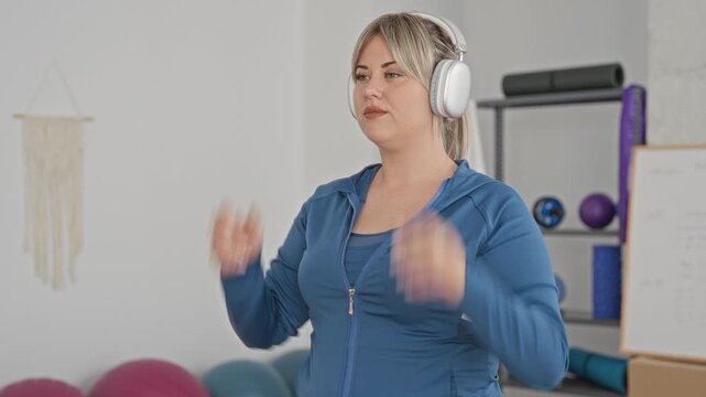 Woman adjusting over ear headphones with hands and closed eyes in a fitness studio among exercise balls and mats; calm focus.