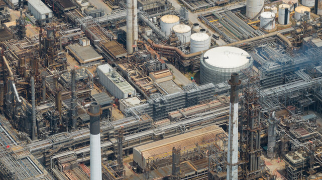 Aerial view of Bangchak Sriracha Refinery with its complex network of industrial pipes, storage tanks, and tall chimneys under bright daylight Si Racha District, Chon Buri, Thailand.
