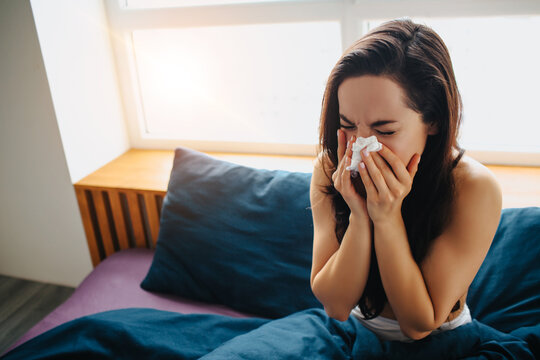 Young beautiful woman in morning bed at home. Blowing nose in white tisue. Flu or caught cold. Suffer from high temperature or virus.