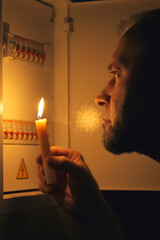 Fototapeta premium Man with Candle Inspecting Electrical Switchboard During Power Outage at Home