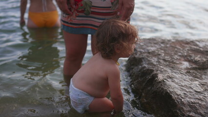 Fototapeta premium Toddler standing in shallow water near large rock while mother stands close, showing parental guidance, safety, and outdoor childhood exploration by the lake