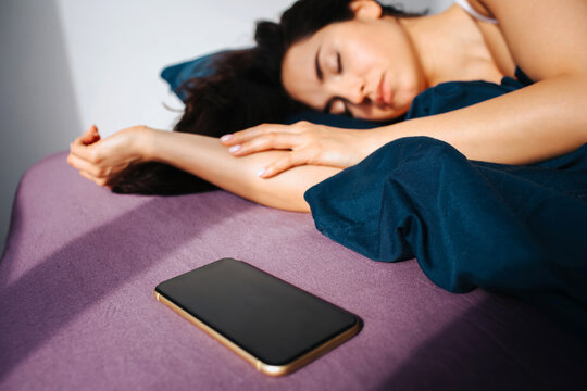 Young beautiful woman in morning bed at home. Sleeping and resting. Sun shines inside. Smartphone lying on ned besides woman.