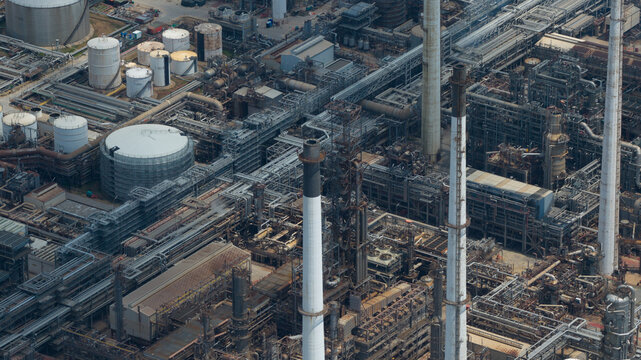Aerial view of the Bangchak Sriracha Refinery featuring a complex network of industrial pipes, storage tanks, and tall chimneys under bright daylight in Si Racha District, Chon Buri, Thailand.