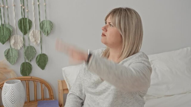 Woman hand mid toss gesture on bed in building, wearing gray sweater and wristwatch, frowning and displeased; annoyance dismissal.