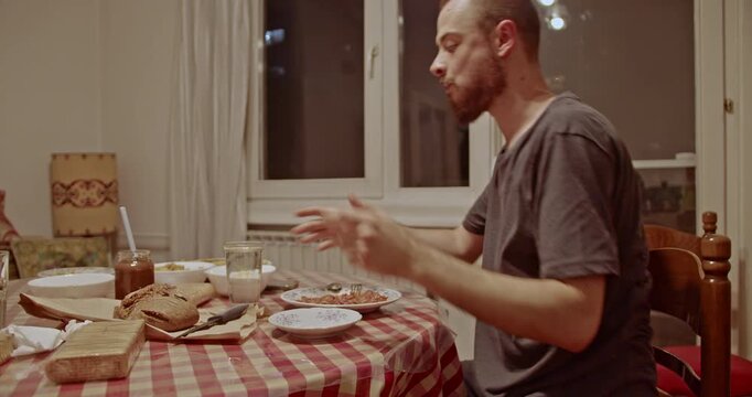 A man with a beard is seen having a casual meal at home. He pours a glass of water and drinks it, enjoying a quiet evening at the dining table with simple food.